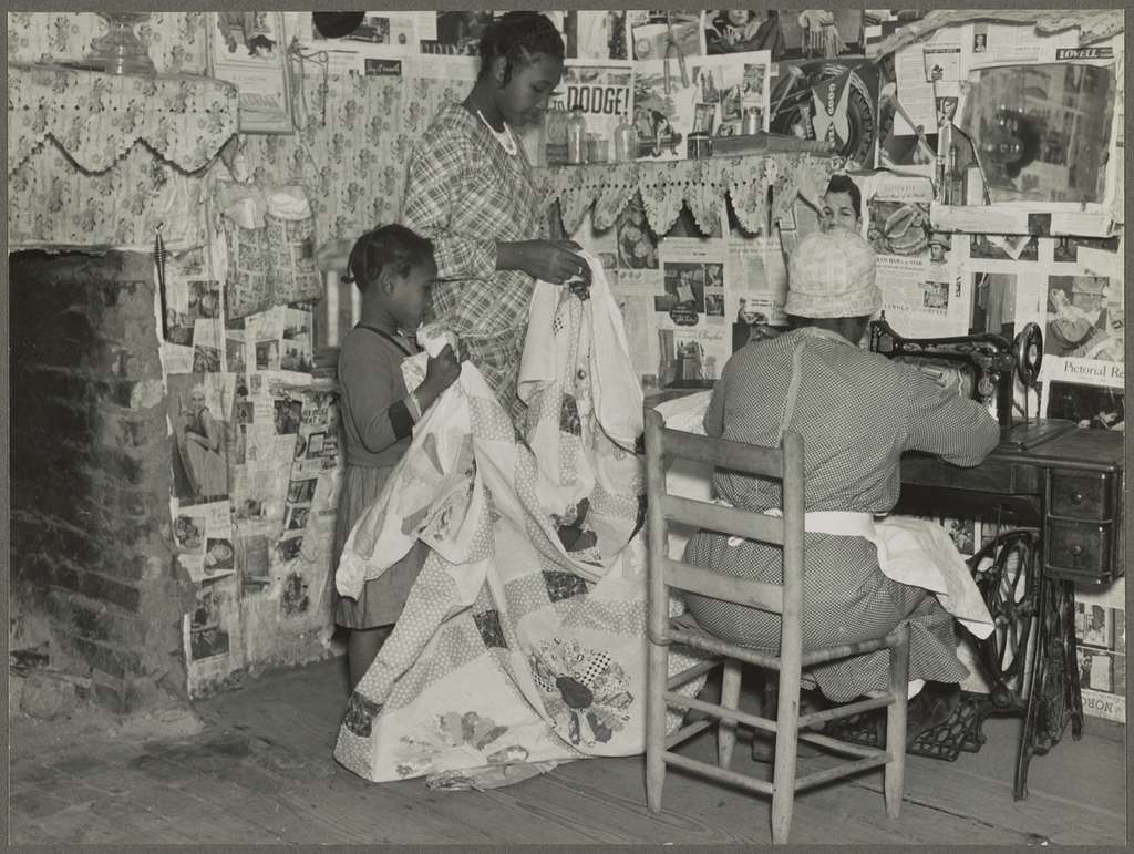 An older Black woman sewing a quilt, while two young children stand by in Gees Bend, Alabama circa 1936.